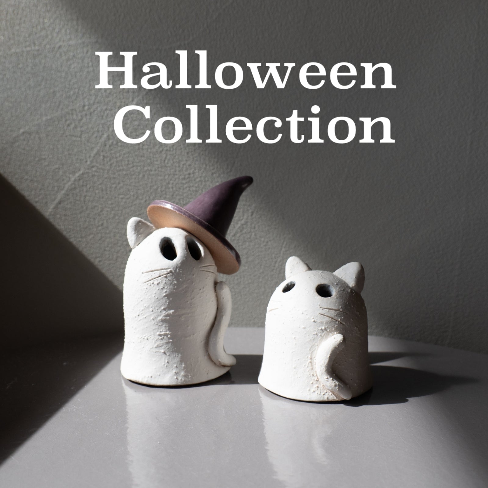 Two white ghost-shaped ceramic figurines on a gray reflective surface against a gray wall, with one ghost wearing a pointed witch hat. Large white text at the top reads 'Halloween Collection'.